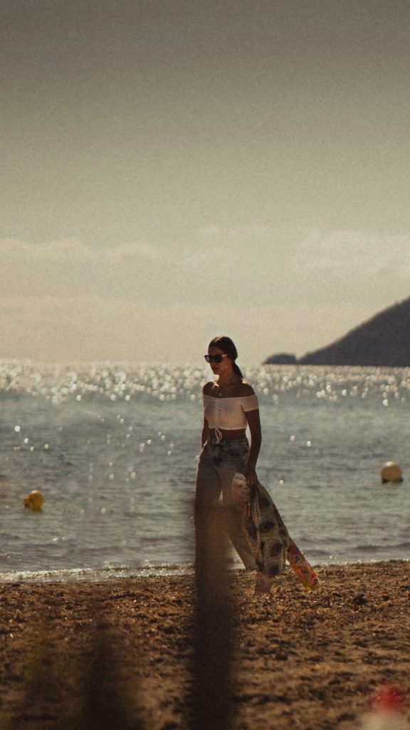 Person walking on a sandy beach near the water’s edge with sunlight reflecting off the sea and distant landforms visible.