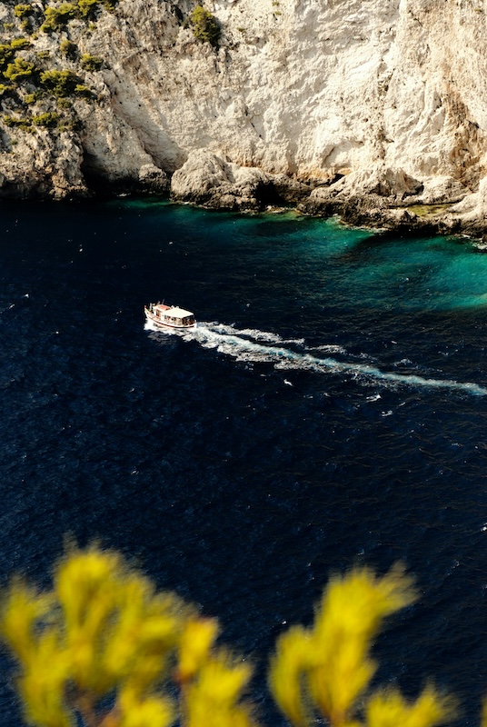 Boat moving through dark blue coastal water with light-colored cliffs and turquoise shoreline in the background.