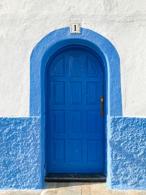 Blue arched wooden door framed by a painted blue arch on a white textured stucco wall, with a number “1” tile positioned above the entrance.