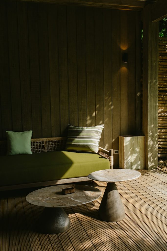 Shaded outdoor lounge area featuring a green cushioned bench, round stone-style tables, wood panel walls, and sunlight filtering through nearby foliage.