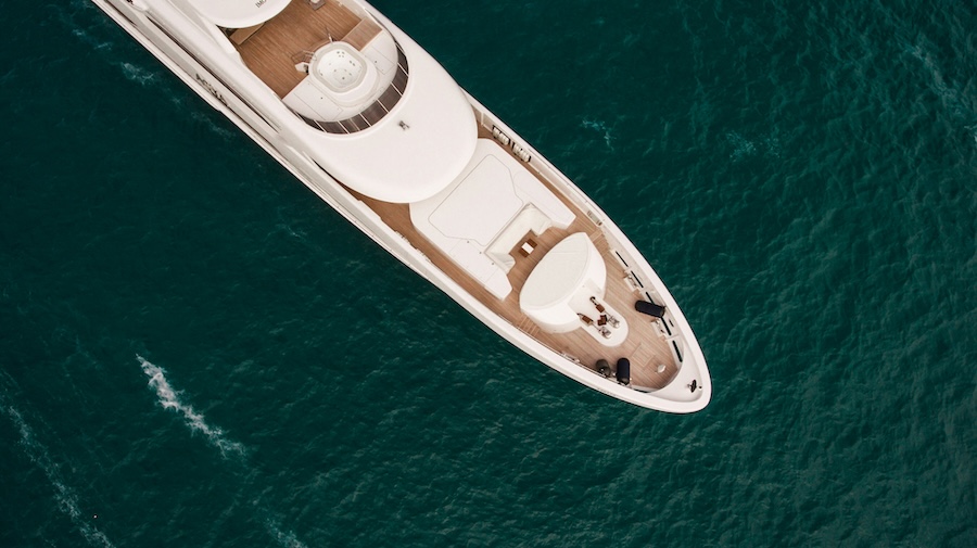Top-down view of a white motor yacht traveling through deep blue water, showing multiple deck levels, wood decking, and a forward lounge area.