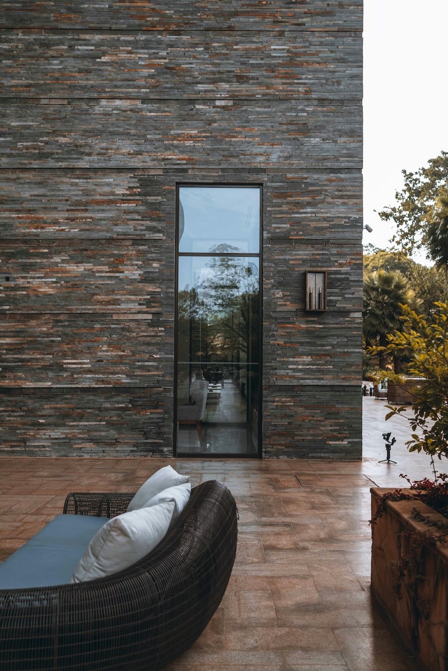 Exterior stone wall with a tall narrow glass door, outdoor patio seating, and surrounding greenery.