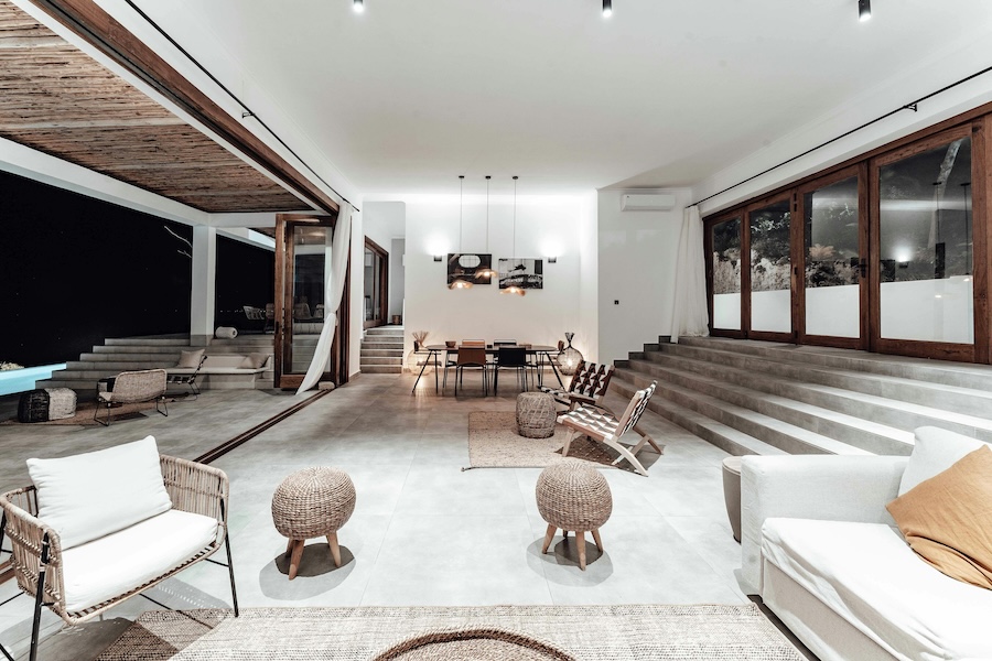 Open indoor–outdoor living area with lounge chairs, woven stools, wide steps, large windows, and a dining table under recessed lighting.