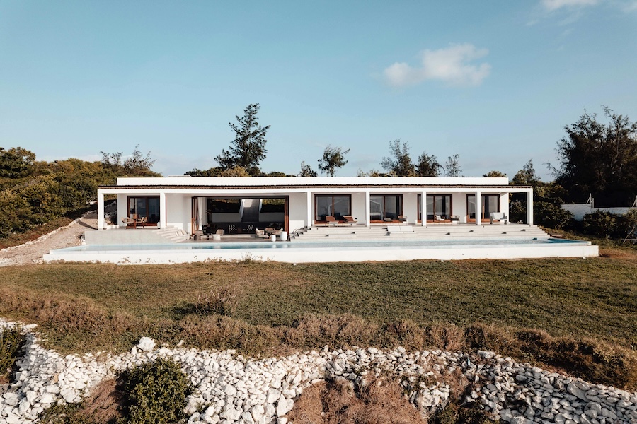 Contemporary single-story white villa with multiple glass-fronted rooms, wide terrace steps, and natural landscape views under a bright sky.