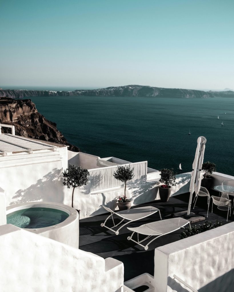 Elevated view of white terraces and potted trees overlooking deep blue water, rocky cliffs, and distant landmasses under a clear sky.