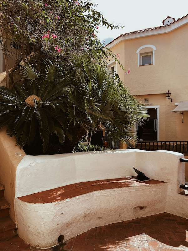 Mediterranean-style courtyard featuring a curved white stucco bench with a tiled seat, surrounded by palm plants and positioned in front of a light-colored building.